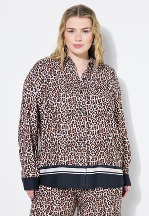 Žena v dlouhé košili s leopardím potiskem a knoflíky, s černobílým pruhovaným lemem, v odpovídajících kalhotách, stojící na bílém pozadí.