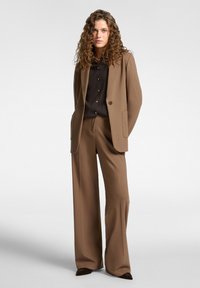 Kvinna med lockigt hår iklädd en brun kostym med vida byxor och en svart skjorta med knappar, stående framför en enkel grå bakgrund.