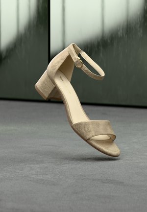 Beige sandal med tekstur og lav blockhæl, med to brede remme og en ankelrem, der lukkes med en guldfarvet spænde.