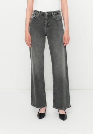 Woman wearing faded black wide-leg jeans, paired with black pointed-toe heels and a white top, standing against a plain white background.