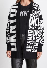 Svart kofta med stora vita "DKNY"-bokstäver i olika riktningar, med ribbat öppet front och texturerad stickad väv. Bärs över en svart skjorta.