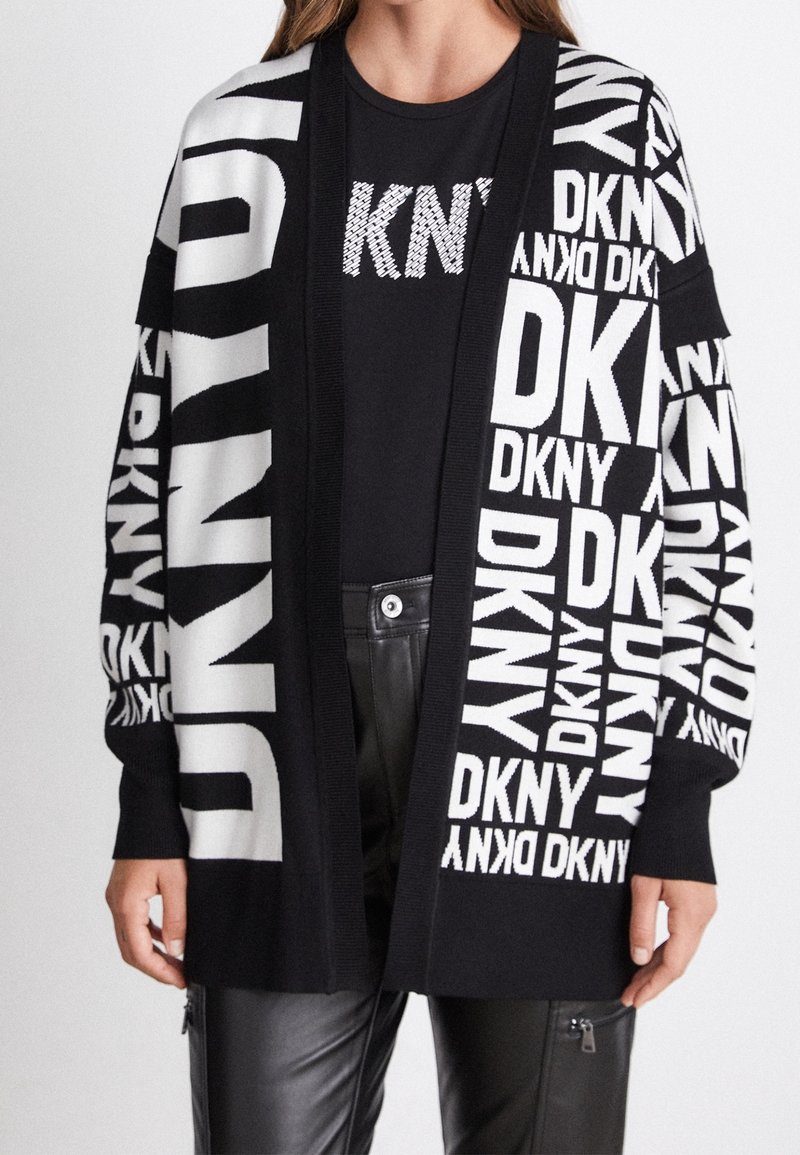 Svart kofta med stora vita "DKNY"-bokstäver i olika riktningar, med ribbat öppet front och texturerad stickad väv. Bärs över en svart skjorta.