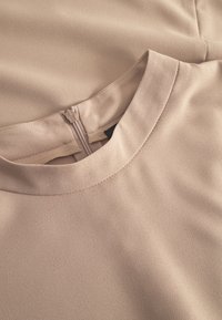 Vaalea beige paita pehmeästä, tekstuurillisesta kankaasta. Omistaa pyöreän kaula-aukon, takana vetoketjun ja reunasauman yksityiskohtia.