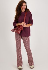 Vrouw in een bordeaux top, met een patroon op uitlopende broek, crème schoenen en een rode gestifte schoudertas, staande tegen een effen achtergrond.