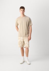 Beige cotton t-shirt with printed design, paired with knee-length light beige camo shorts and white sneakers. Simple, casual outfit.