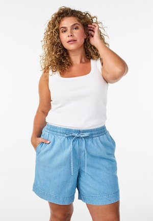 Lockige Frau trägt ein weißes Tanktop und hellblaue Jeans-Shorts mit Kordelzug, steht mit einer Hand in der Tasche und berührt mit der anderen ihr Haar.