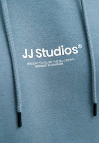 Sweat à capuche bleu clair en tissu doux, doté d'une capuche à cordon, avec le logo "JJ Studios" et un texte supplémentaire imprimé en blanc.