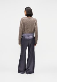 Γυναίκα που φαίνεται από πίσω, φοράει καφέ χνουδωτό πουλόβερ και χαλαρά σκούρα μωβ σατέν παντελόνια μπροστά σε απλό ανοιχτό φόντο.