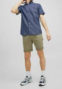 Džínsová košile s krátkými rukávy a límečkem na knoflíky, doplněná o vykasané khaki kraťasy. Osoba má na sobě bílé ponožky a sportovní boty.