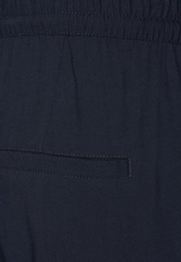 Navyblauwe stof met een gladde textuur, voorzien van een platte zak en een elastische tailleband. De stiksels zijn subtiel en matchen bij de kleur van de stof.