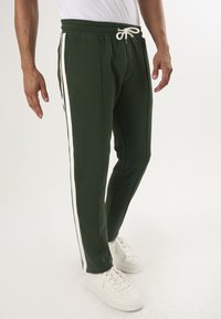 Gröna sweatshirtbyxor med vita sidostråkar, dragsko i midjan och avslappnad passform, i kombination med vita sneakers på en neutral bakgrund.