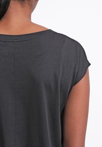 Person som bär en kortärmad svart skjorta, visad bakifrån med en synlig överarm och axel mot en enfärgad bakgrund.