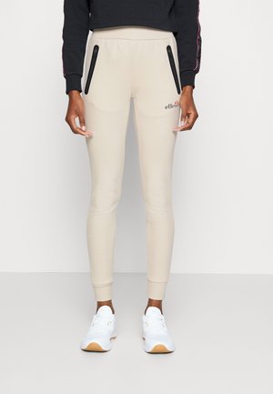 Person iført beige joggers med sorte lynlåslommer, sort cropped sweatshirt og hvide sneakers, stående mod en ensfarvet baggrund.