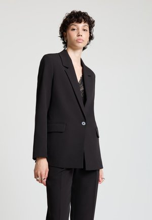 Femme aux cheveux courts et bouclés, portant un blazer noir ajusté et un pantalon assorti, debout devant un fond gris clair uni.