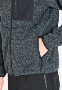 Giacca in pile grigia con tasca sul petto con zip, tasche laterali con zip e tessuto testurizzato. Accentuata da rifiniture e cuciture nere.