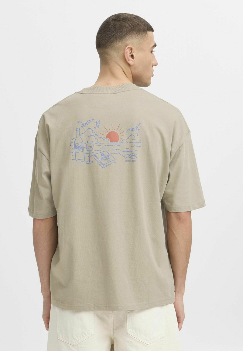 T-shirt à manches courtes kaki avec un graphique coloré au dos représentant un coucher de soleil, une bouteille de vin, un verre et des oiseaux dans une scène de plage.