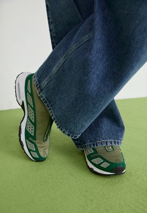 Sneakers verts et beiges avec des panneaux en mesh, forme arrondie, semelle en caoutchouc et accents perforés, portés avec un jean en denim à jambes larges.