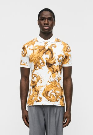 Férfi fehér galléros pólóban, díszes arany és barna barokk stílusú hullámmintákkal, szürke nadrágban, sima háttér előtt állva.