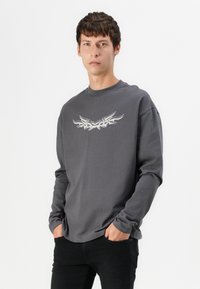 Un tânăr cu părul scurt și brunet poartă un tricou cu mâneci lungi de culoare gri închis, cu un design tribal deschis, pantaloni negri, având mâinile în buzunare.