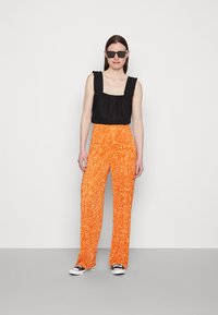 Top de alças com decote quadrado preto e alças com franzidos, combinado com calças largas laranja com um padrão em espiral e ténis preto e branco.