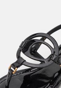 Sorte patentsandaler med et cirkulært hardware-element, slanke stropper og en glat, blank overflade. Detalje med guldfarvet spænde inkluderet.