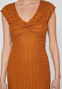 Braunes Strickkleid mit tiefem V-Ausschnitt und kurzen Ärmeln, ausgestattet mit einem strukturierten Muster, das Schuppen ähnelt, sowie einer Knotendetaillierung am Bustbereich.