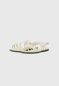 Beige flate sandaler med flere tynne stropper og ankelspenne på svart såle, vist side om side på hvit bakgrunn.