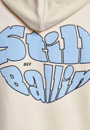 Cremefarvet hættejakke med store blå bogstaver, der står "Still Ballin" i en grafisk stil. Blødt bomuldsstof med en glat tekstur.