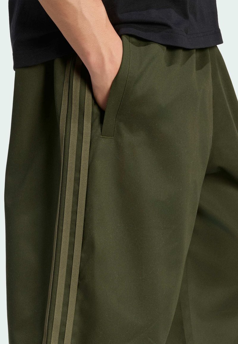 Mano descansando en el bolsillo de unos pantalones verde oliva con tres rayas verticales a lo largo del lateral, combinados con una camisa negra.