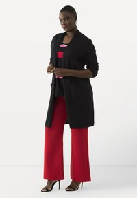 Schwarzer, langer Cardigan über einem mehrfarbig gestreiften Oberteil und roten weit geschnittenen Hosen. Mit Taschen, glattem Stoff und Absatzsandalen.