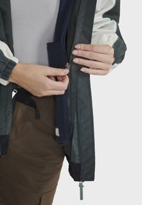 Veste imperméable avec un mélange de sections vert foncé et blanches, dotée de poignets élastiques et d'une fermeture éclair à l'avant avec une doublure marine.