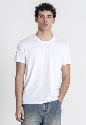 Joven con pelo rizado de pie mirando hacia adelante, lleva una camiseta blanca lisa y vaqueros azules desgastados, una mano en el bolsillo.