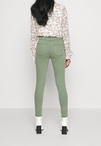 Jeans skinny de tiro alto verdes con una textura suave y dos bolsillos traseros, combinados con una blusa de manga larga con estampado floral y botas blancas de tacón grueso.