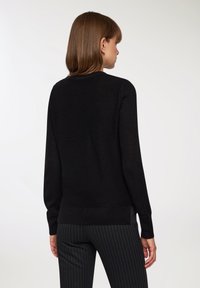 Maglione nero a maglia con scollo rotondo, maniche lunghe e orlo a costine; texture liscia e design minimalista, indossato su pantaloni a righe.