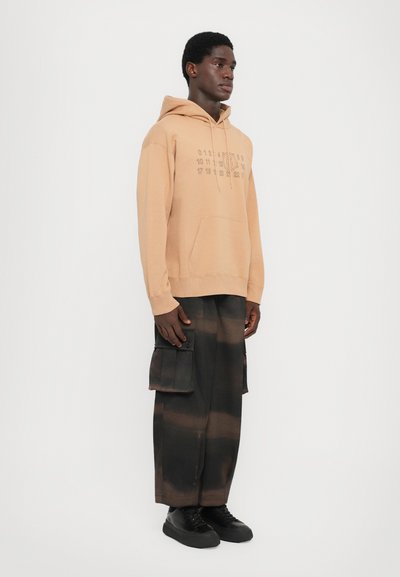 Ljusbrun hoodie med en framficka och tryckta siffror; kombinerad med svarta tie-dye cargo-byxor med sidfickor. Svarta skor fulländar looken.