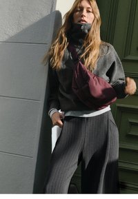 Maglione grigio a righe con colletto alto e borsa a tracolla bordeaux, abbinato a pantaloni grigi a righe larghe, sullo sfondo di un muro testurizzato.