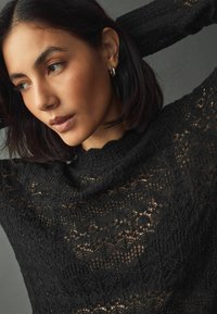 Maglione in pizzo nero con un motivo testurizzato, colletto a coste e maniche lunghe. Il tessuto è leggero e trasparente, rivelando la pelle sottostante.