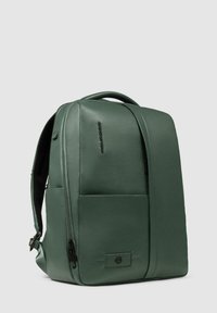 Zaino verde in pelle vegana con forma strutturata, tasca frontale con zip, manico superiore e spallacci regolabili. Texture liscia, design minimale.