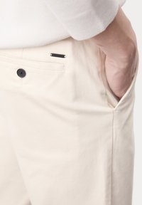 Beige broek van lichtgewicht stof, met een voorkantoor, zwart knoopsluiting en een metalen merklabel op de tailleband.