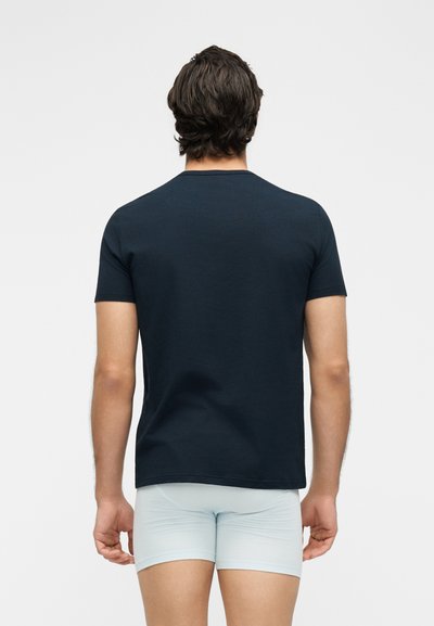 Άνδρας μοντέλο με την πλάτη γυρισμένη, φοράει σκούρο μπλε κοντομάνικο πουκάμισο και ανοιχτόχρωμα εφαρμοστά σορτς σε απλό φόντο.