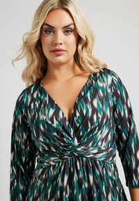 Abito a portafoglio in nero, verde mare e bianco con un motivo astratto; presenta una profonda scollatura a V, maniche lunghe e un dettagliato nodo in vita. Tessuto dalla texture liscia.