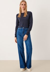 Granatowy sweter, ściągacze na mankietach; błękitne spodnie o wysokim stanie, szerokie nogawki; beżowe buty na platformie z teksturowanymi bokami; neutralne tło.