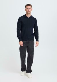 Dunkelblauer Pullover mit spitzem Kragen, geripptem Saum und langen Ärmeln, kombiniert mit schwarzen Jeans und weißen Sneakers.