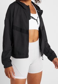 Sweat à capuche zippé noir en tissu doux, avec une capuche doublée et des accents en maille. Associé à un brassière de sport blanche et un short clair.