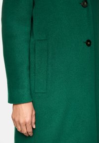 Der grüne Wollmantel zeichnet sich durch eine glatte Textur, eine Knopfleiste und seitliche Eingrifftaschen aus. Der Ärmel hat eine leichte Glockenform, was das strukturierte Design zusätzlich betont.