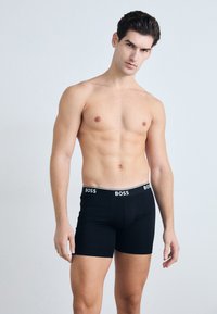 Schwarze Boxershorts mit enger Passform, ausgestattet mit einem weißen Bund, der mit "BOSS" versehen ist. Das Material wirkt glatt und elastisch, ausgelegt für Komfort.