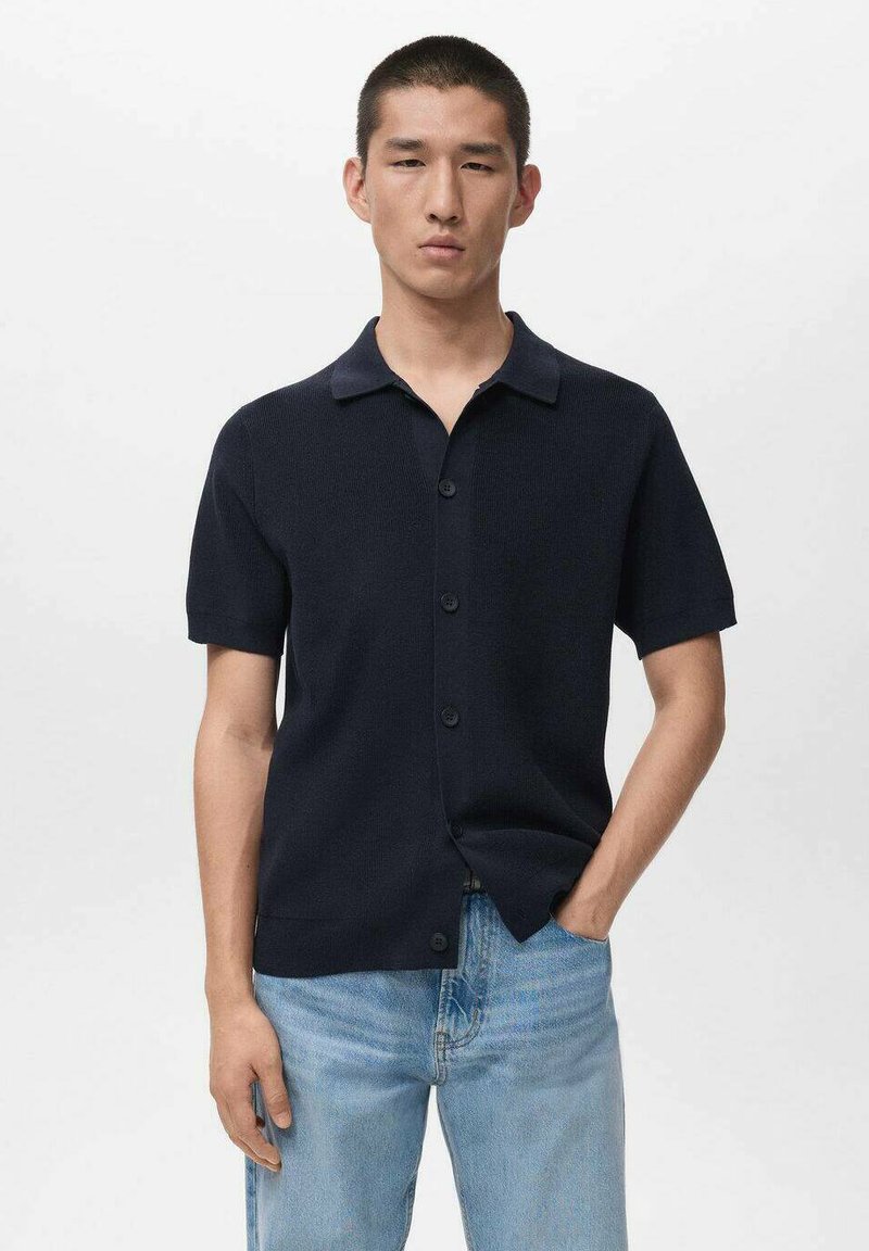Maglietta a maniche corte in maglia blu navy con collo a camicia, chiusura frontale con bottoni e tessuto testurizzato. Abbinata a jeans blu chiaro.