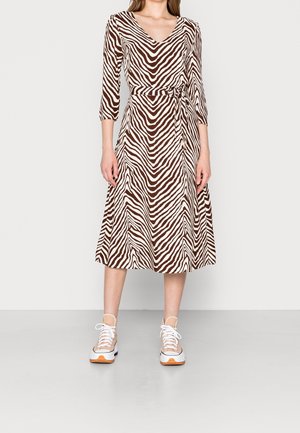 Vrouw draagt een midi-jurk met bruin-witte zebraprint, 3/4 mouwen en een strik in de taille, gecombineerd met witte en beige sneakers.
