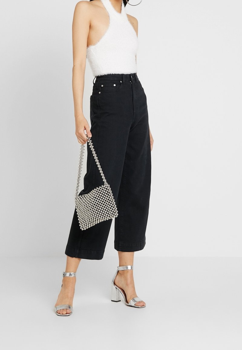 Femme portant un haut à fines bretelles en tissu duveteux blanc, un pantalon noir taille haute à jambes larges, des sandales à talons blocs argentés, et tenant un sac à main argenté orné de perles.