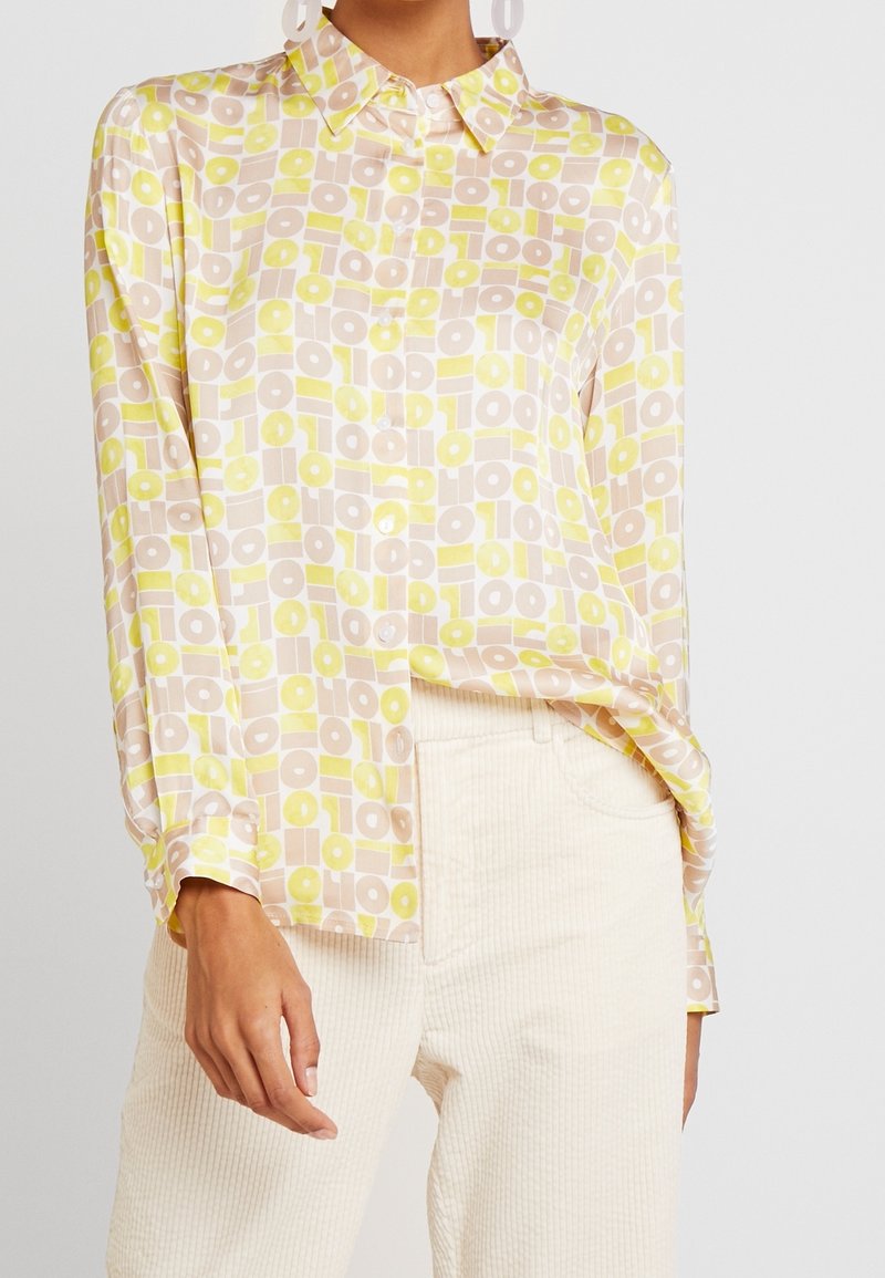 Femme portant une chemise à manches longues avec un motif géométrique beige et jaune, associée à un pantalon en velours côtelé écru.